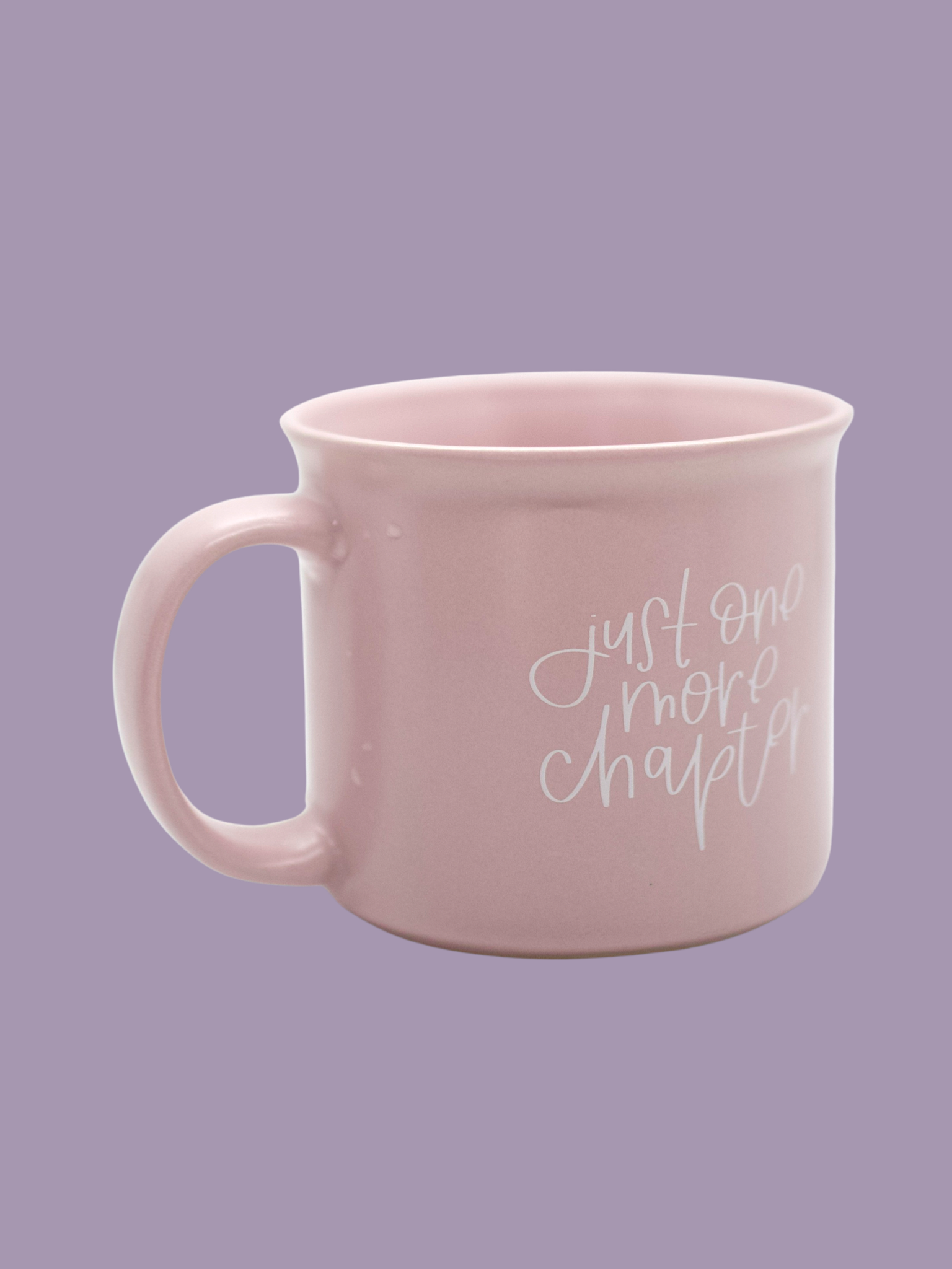 A matte lilac ceramic mug with the phrase 'Just One More Chapter' printed in white on the side, held against a purple background.