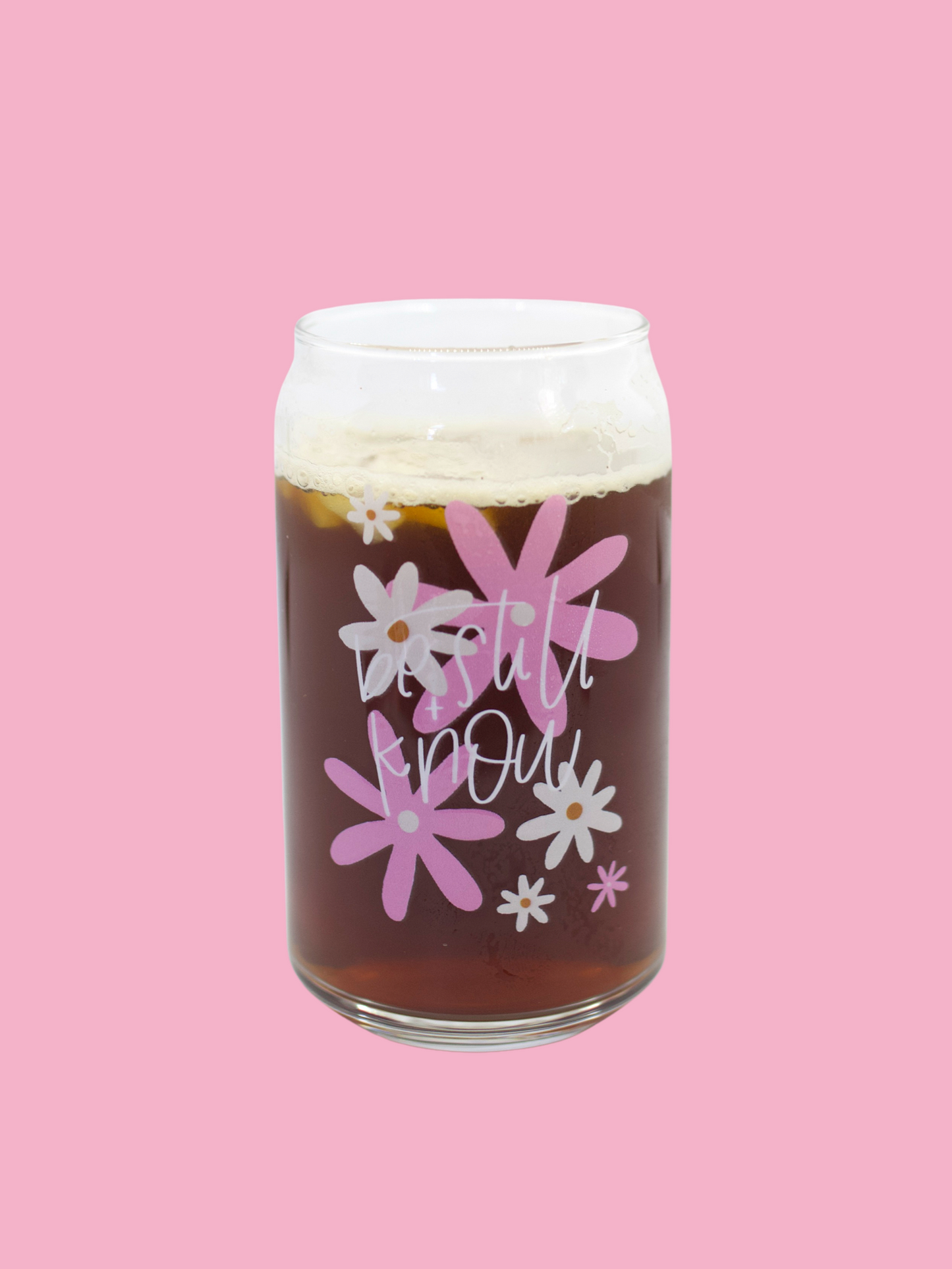 A clear can-shaped glass with pink and ivory daisy designs and the text 'be still and know' on it.