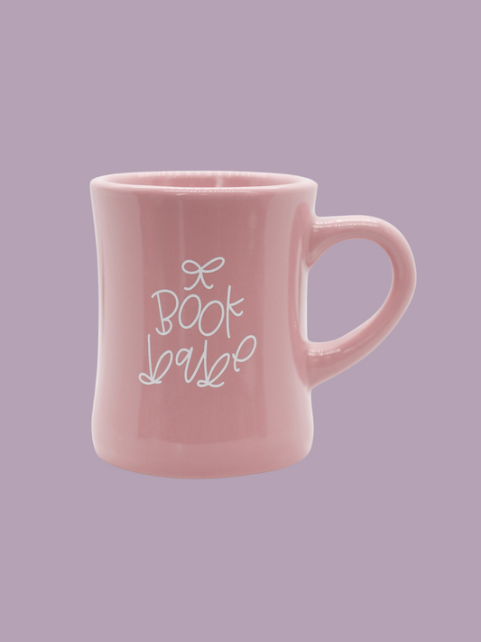 A glossy pink ceramic mug with 'book babe' printed in white hand-lettering and a small bow detail.