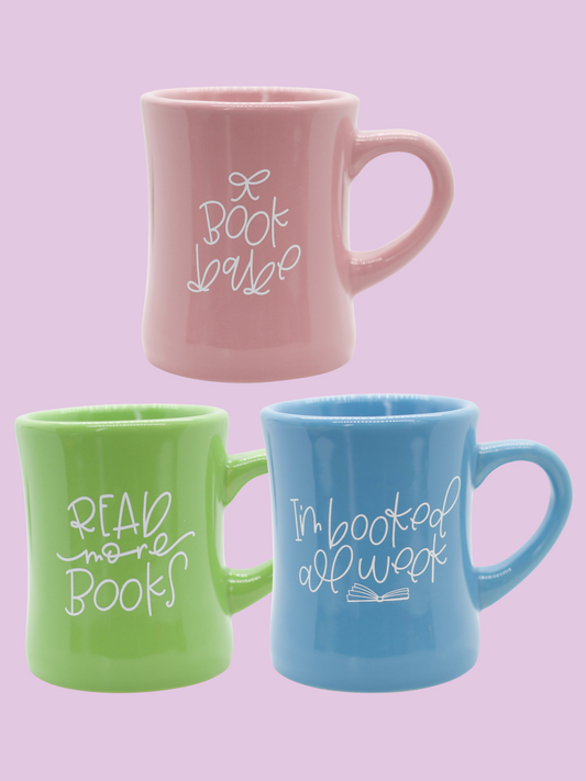 Set of three ceramic mugs in pink, blue, and green colors with text and book-related imagery on them.