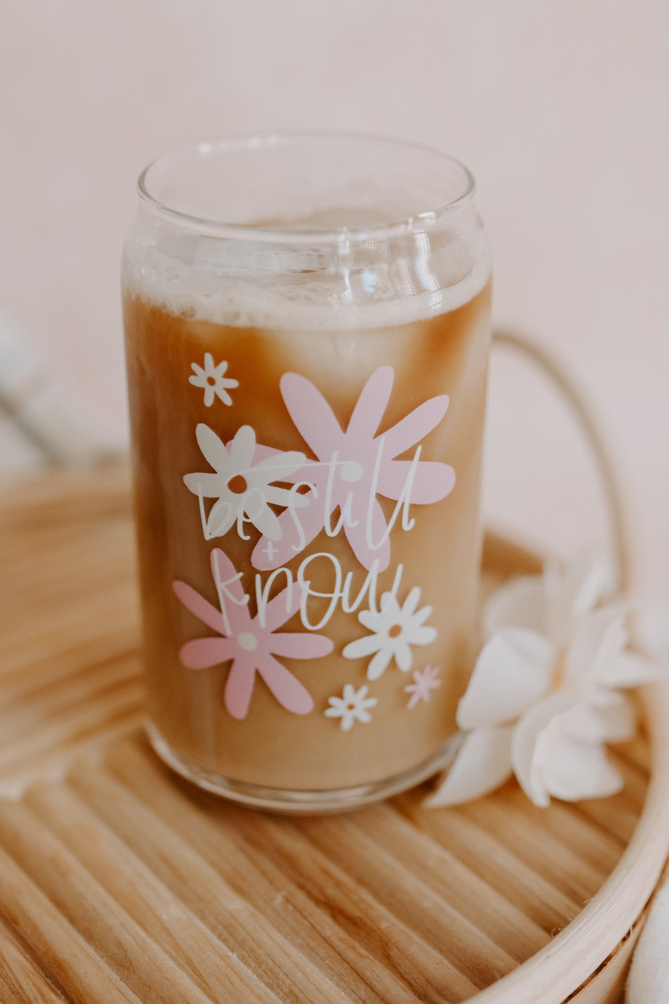 A clear can-shaped glass with pink and ivory daisy designs and the text 'be still and know' on it.