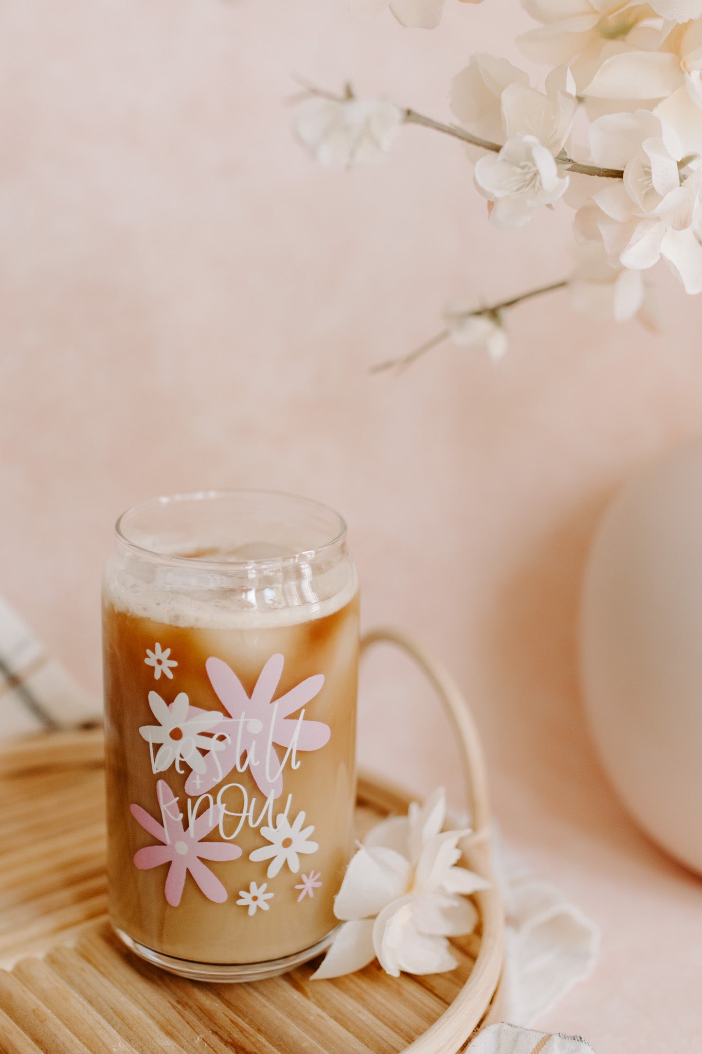 A clear can-shaped glass with pink and ivory daisy designs and the text 'be still and know' on it.