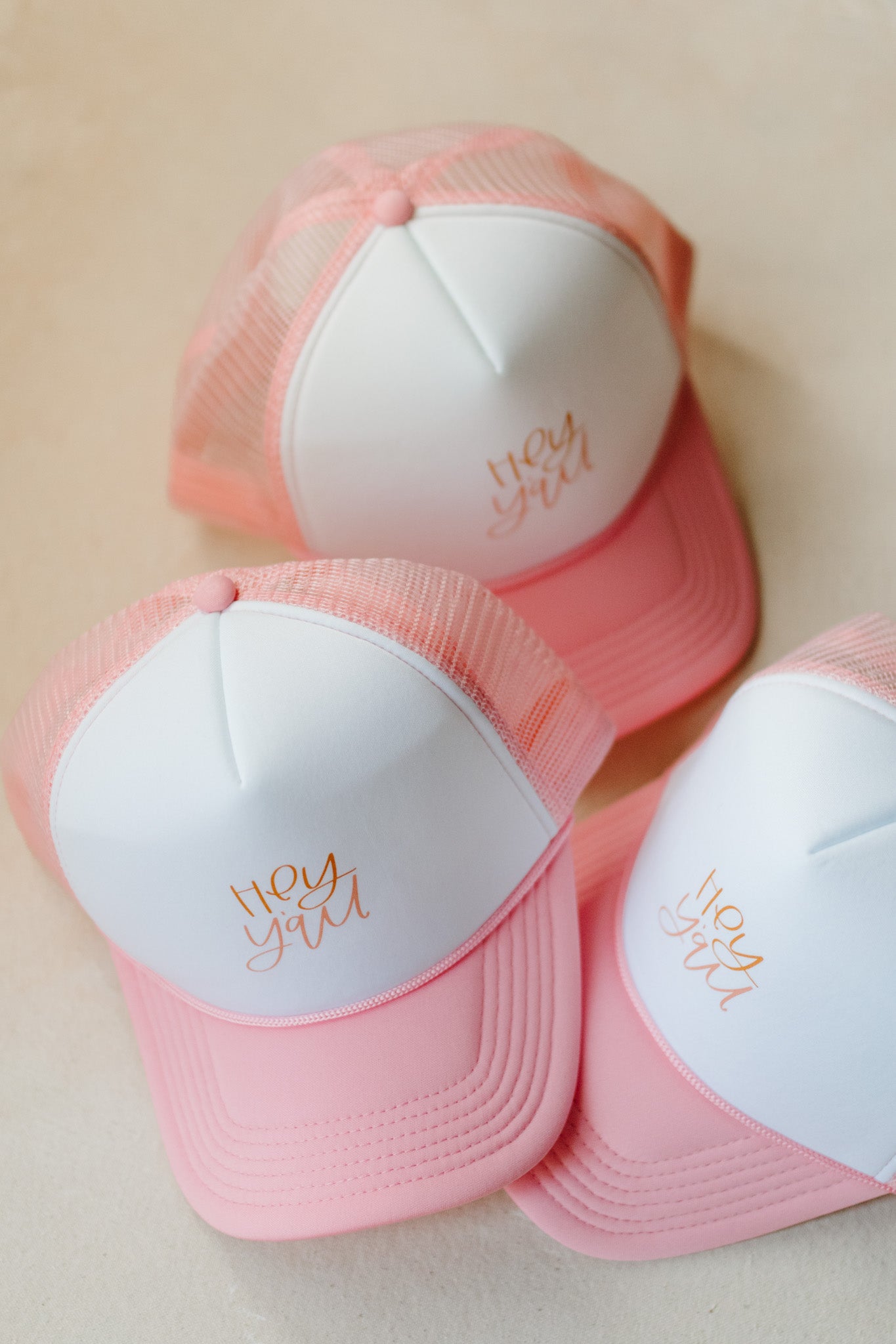 A pink and white trucker hat with 'hey y'all' text in gold and pink on the front.