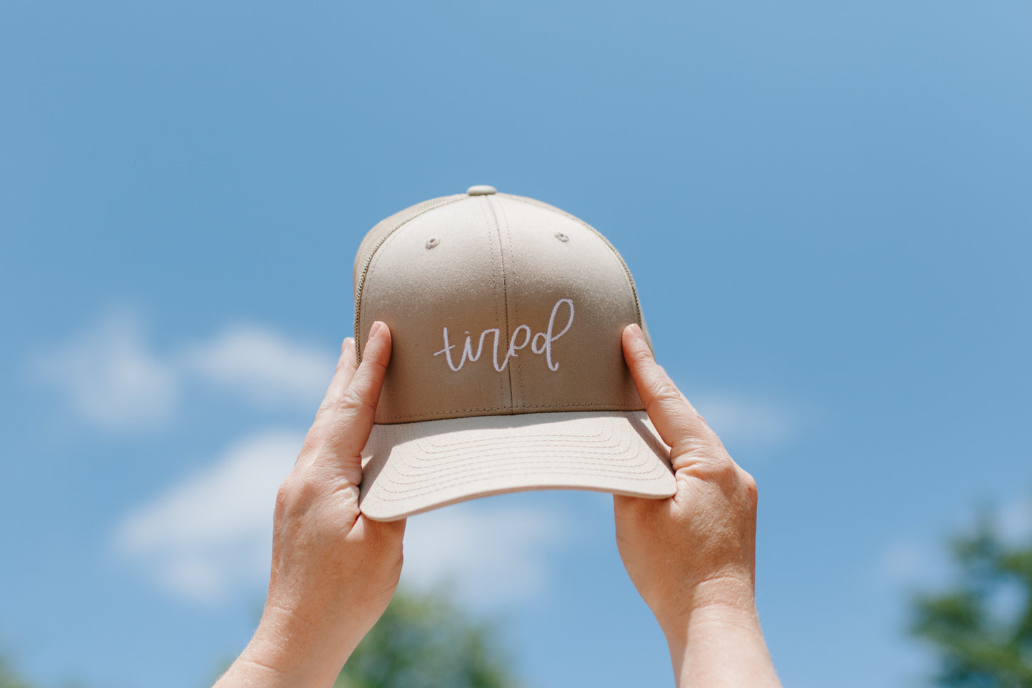 A beige embroidered retro trucker hat with the word 'tired' stitched in white thread on the front.