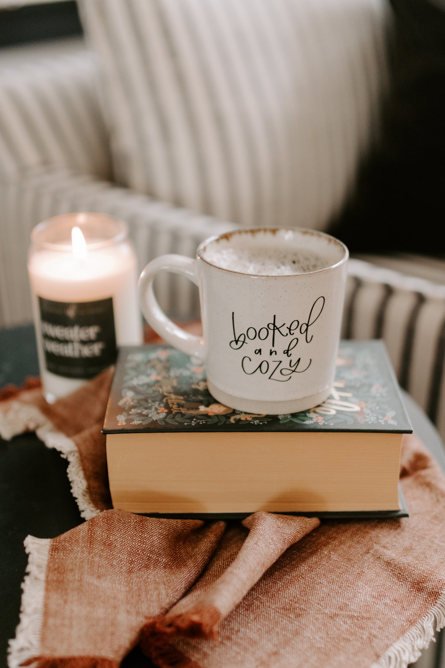 Mug with 'booked and cozy' text on a book next to a lit candle on a blanket.
