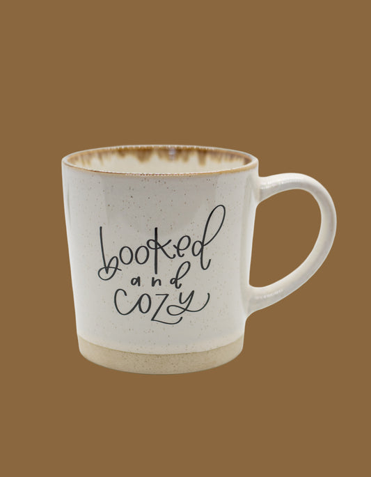 White mug with 'booked and cozy' text on a brown background